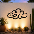 Black metal cloud wall art with flowing silhouette cutouts, mounted on a textured outdoor wall above a desert garden with cacti and warm uplighting.