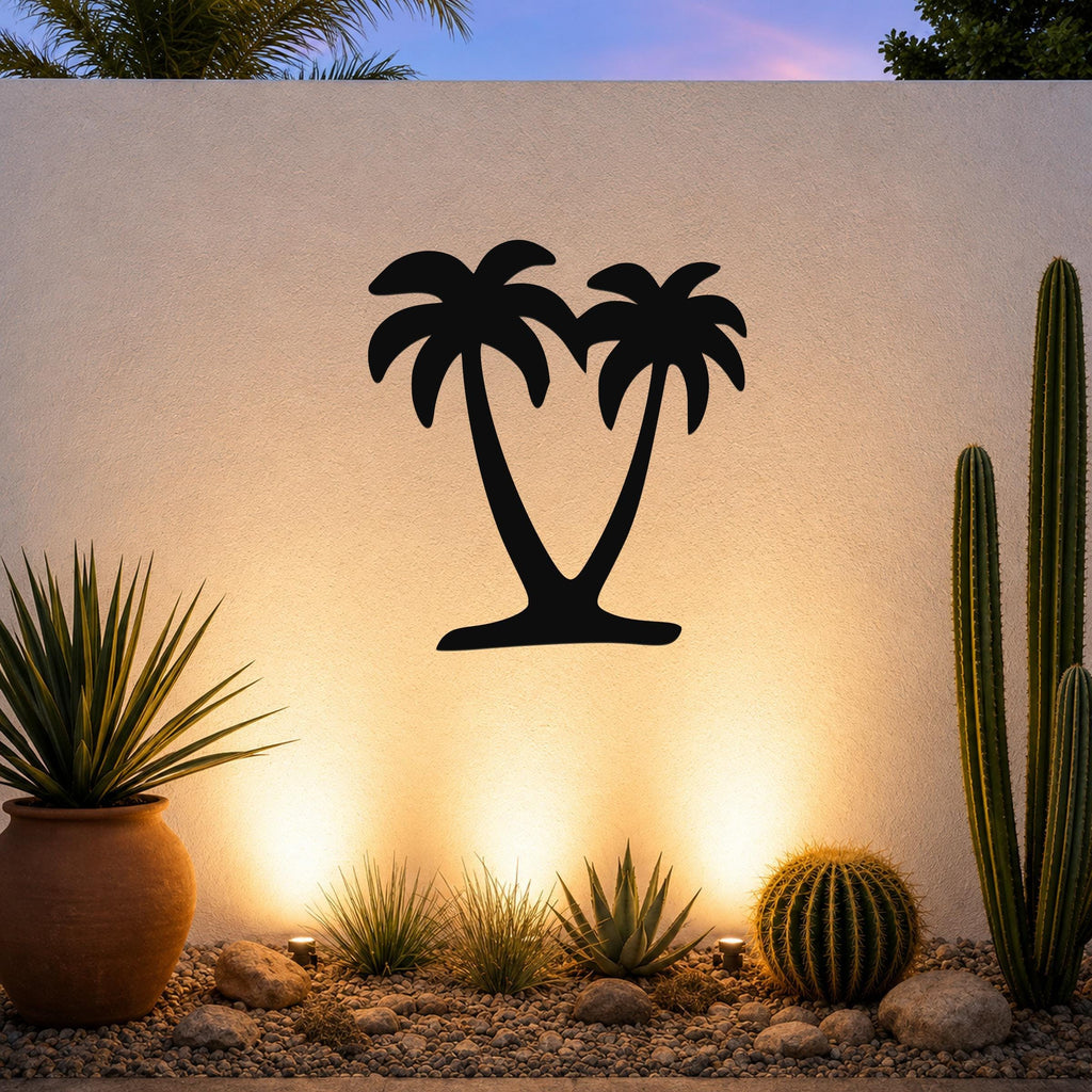 Black metal palm tree wall art with two silhouetted trunks and fronds, mounted on a white outdoor wall above a desert garden with cacti and warm uplighting.