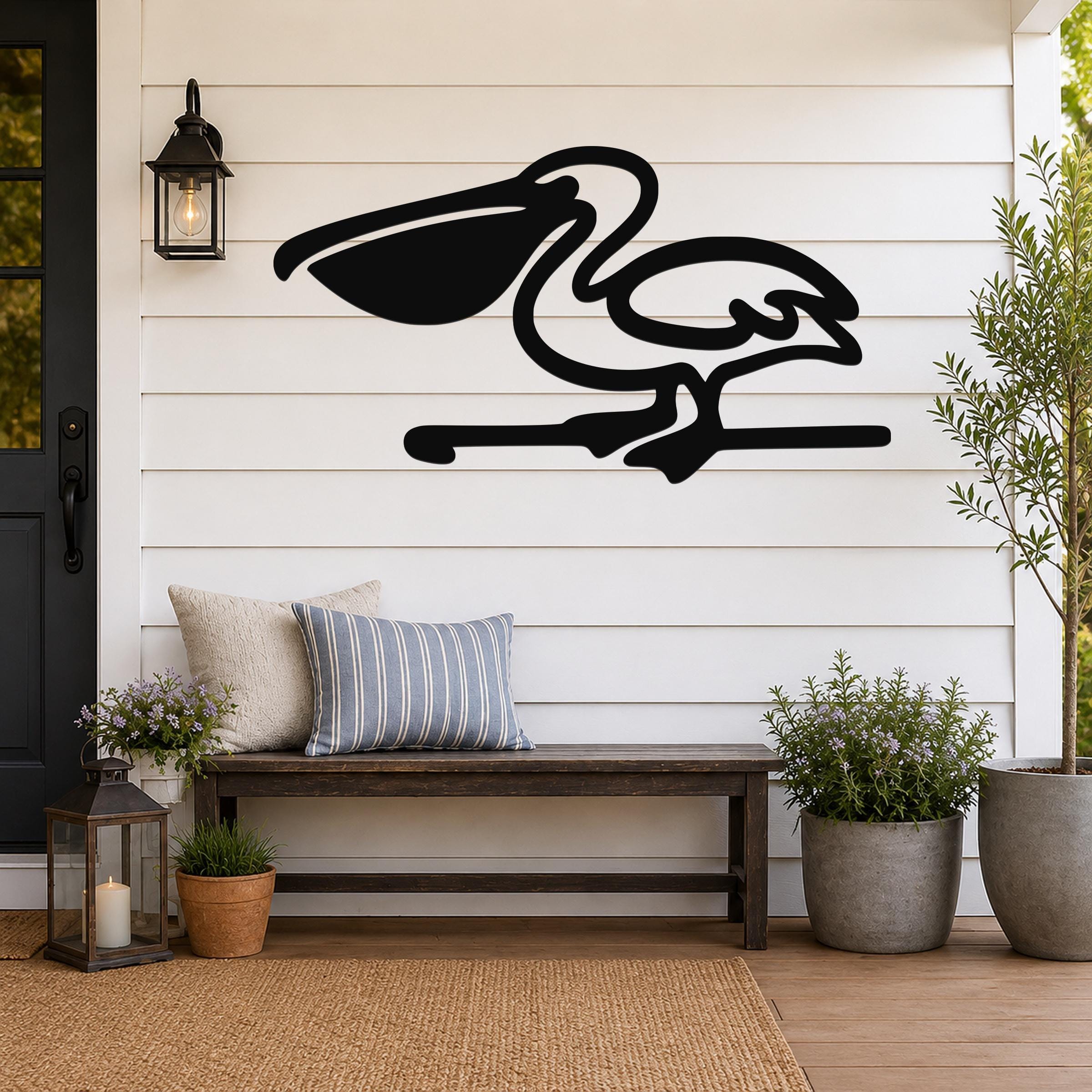Black metal pelican wall art in silhouette style, mounted on a white shiplap wall above a wooden bench on a porch.