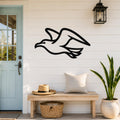 Black metal seagull wall art with a silhouette design, mounted on a white shiplap wall above a wooden bench in a coastal entryway.
