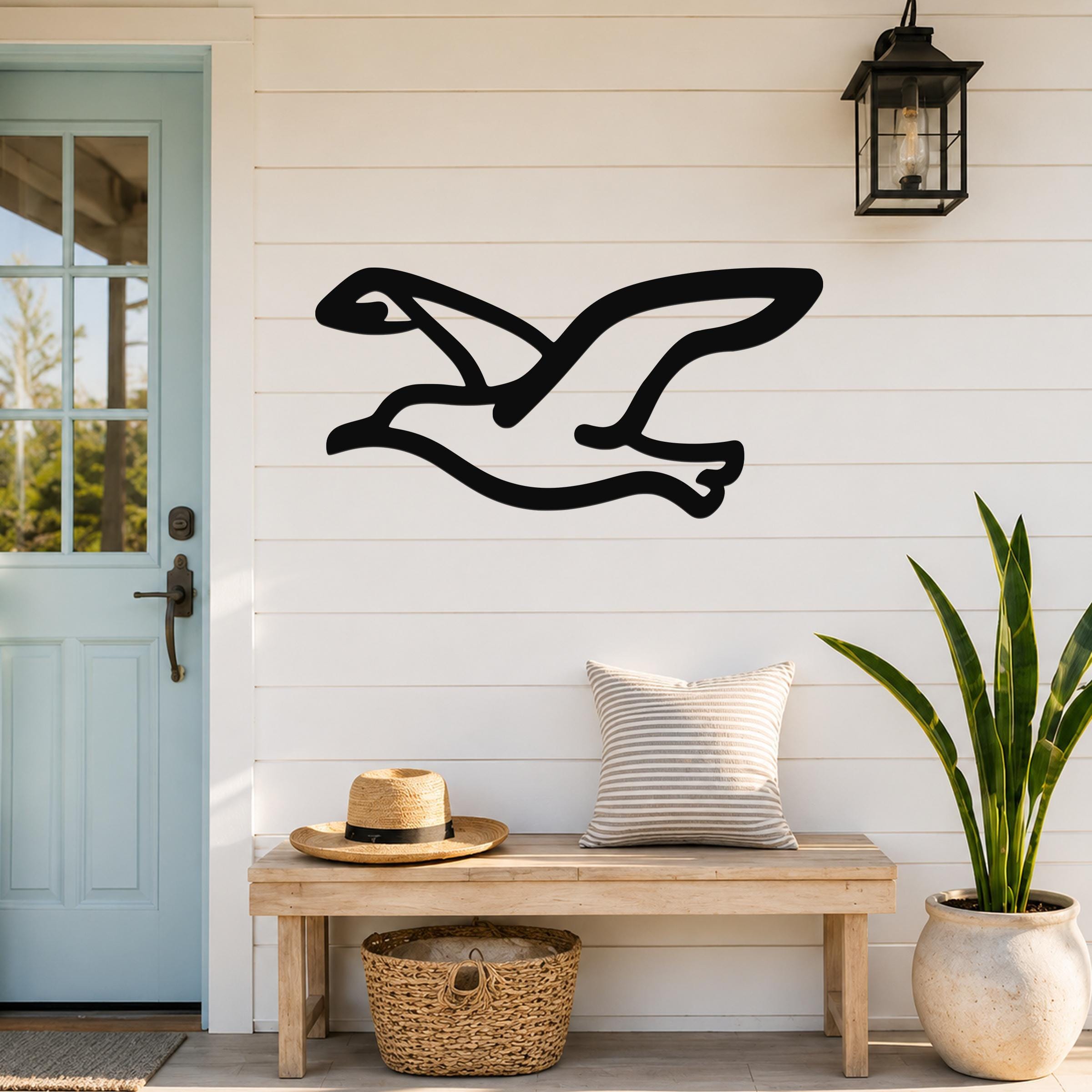 Black metal seagull wall art with flowing wing cutouts, mounted on a white shiplap wall above a wooden bench in a coastal entryway.