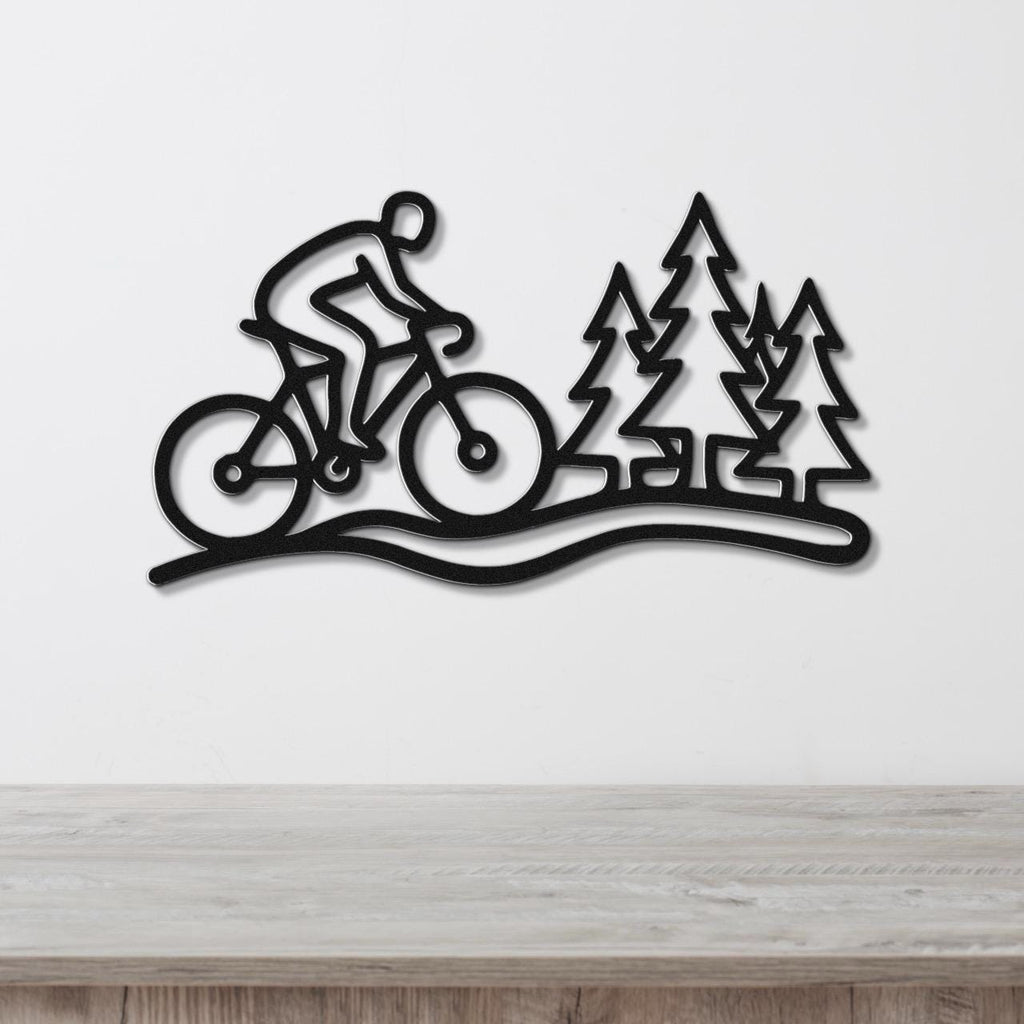 Black metal wall art featuring a cyclist silhouette on a trail with pine tree cutouts, mounted on a white wall above a wooden surface.