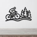 Black metal wall art featuring a cyclist silhouette on a trail with pine tree cutouts, mounted on a white wall above a wooden surface.