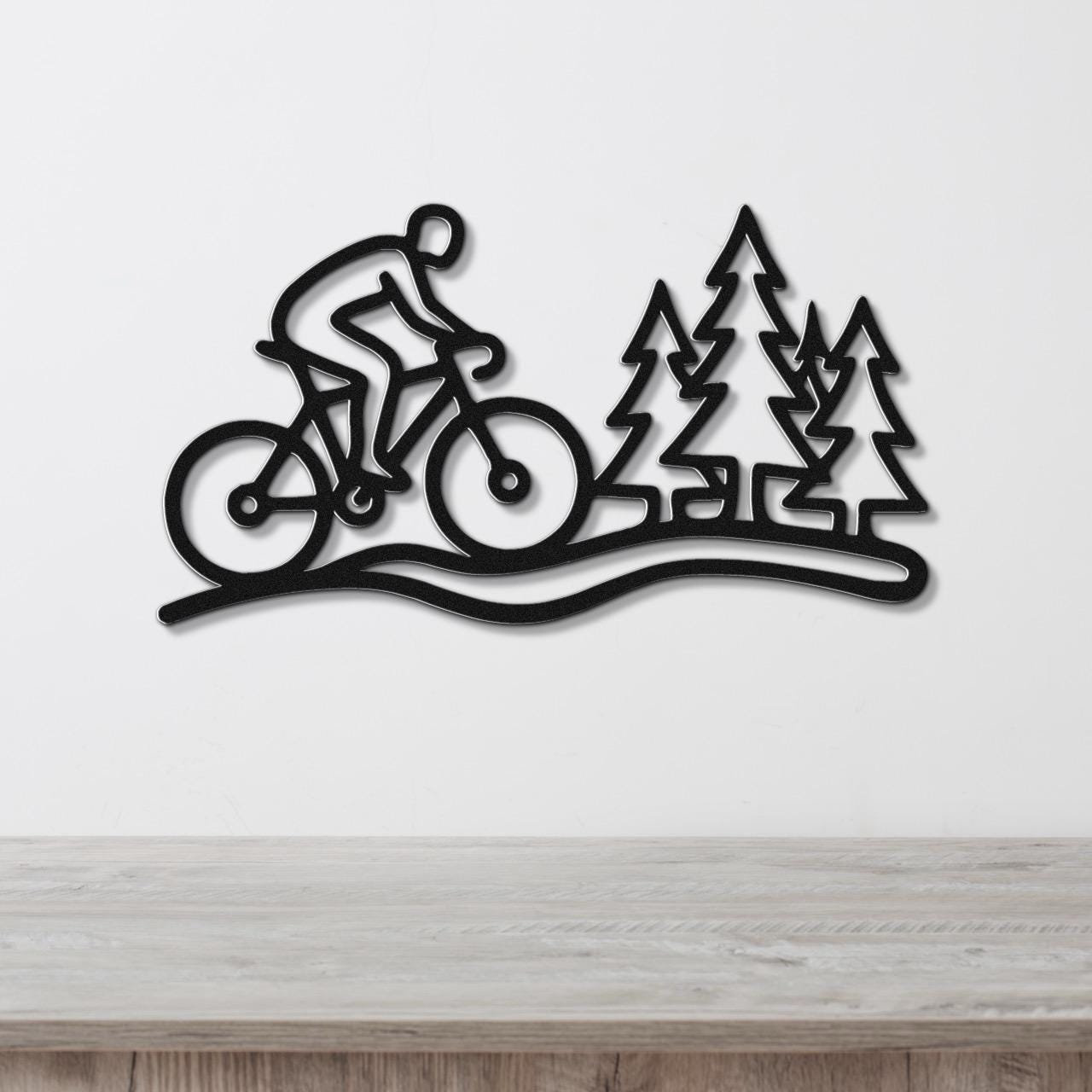 Black metal wall art featuring a cyclist silhouette on a trail with pine tree cutouts, mounted on a white wall above a wooden surface.