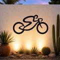 Black metal bicycle wall art with a bold silhouette design, mounted on an outdoor wall above a desert garden with cacti and warm uplighting.