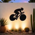 Black metal wall art featuring a bold silhouette of a cyclist in motion, mounted on an outdoor wall above a desert garden with cacti and warm uplighting.