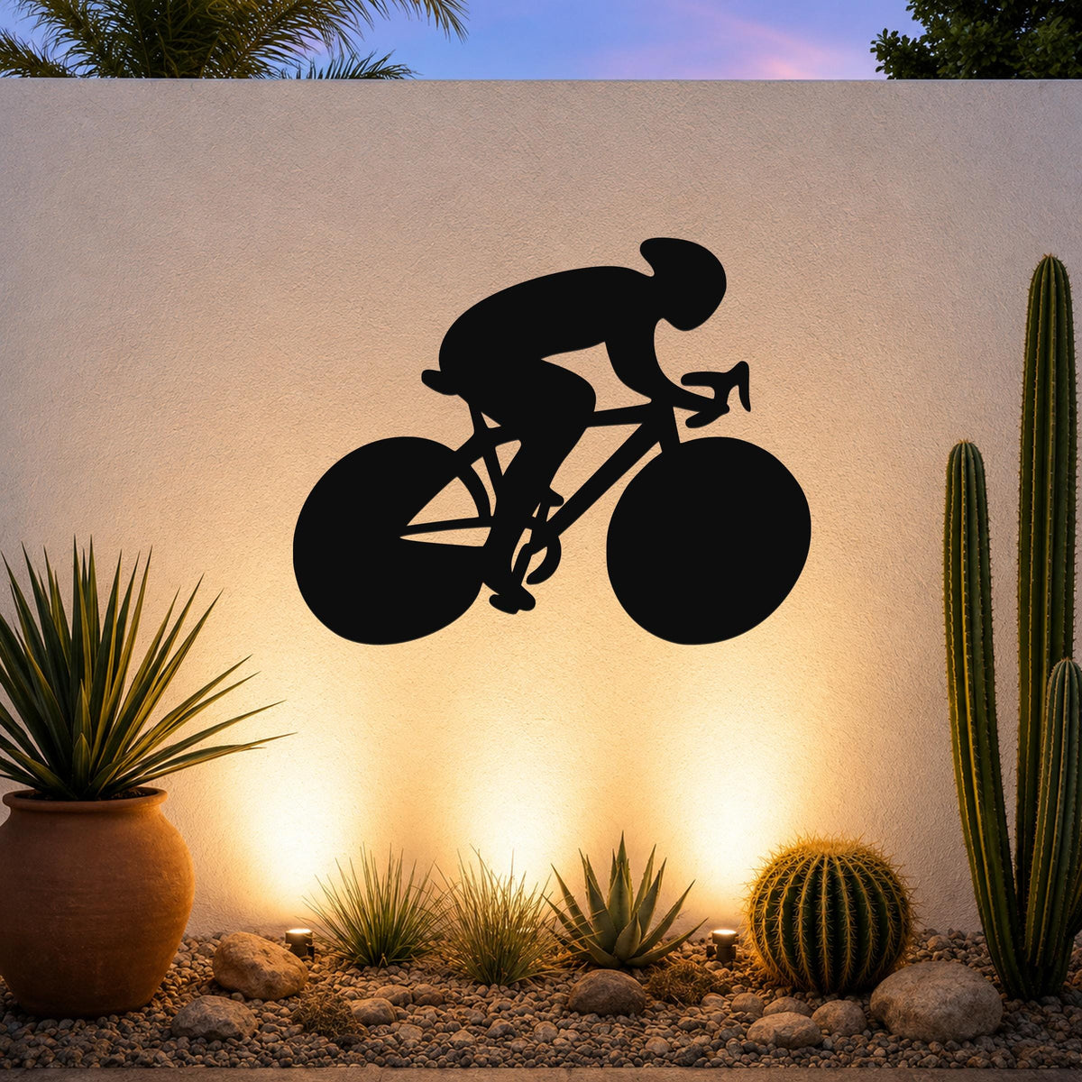 Black metal wall art featuring a bold silhouette of a cyclist in motion, mounted on an outdoor wall above a desert garden with cacti and warm uplighting.