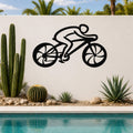 Black metal wall art of a cyclist silhouette with cutout wheels, mounted on a white outdoor wall above a pool with cacti and palm trees.