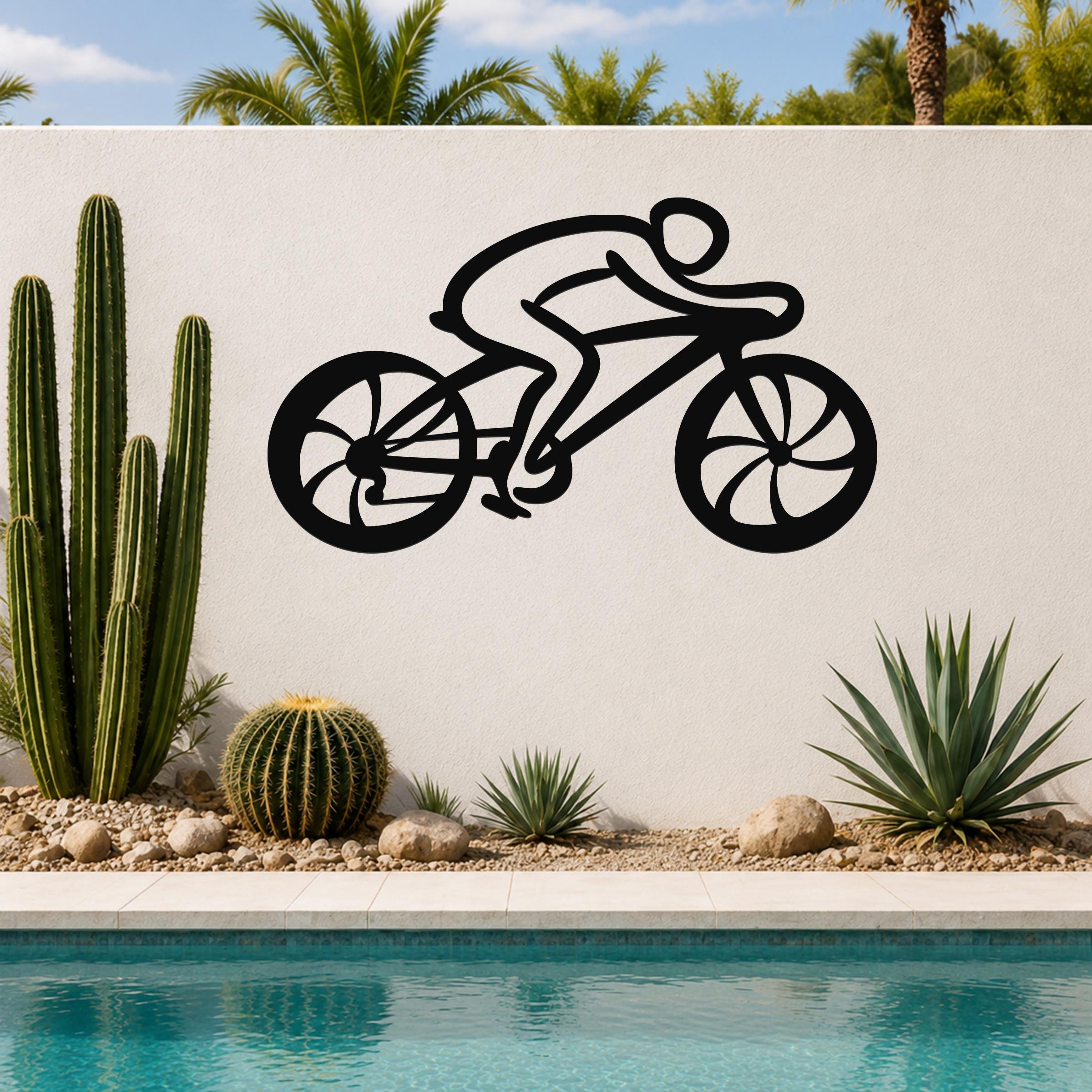 Black metal wall art of a cyclist silhouette with cutout wheels, mounted on a white outdoor wall above a pool with cacti and palm trees.