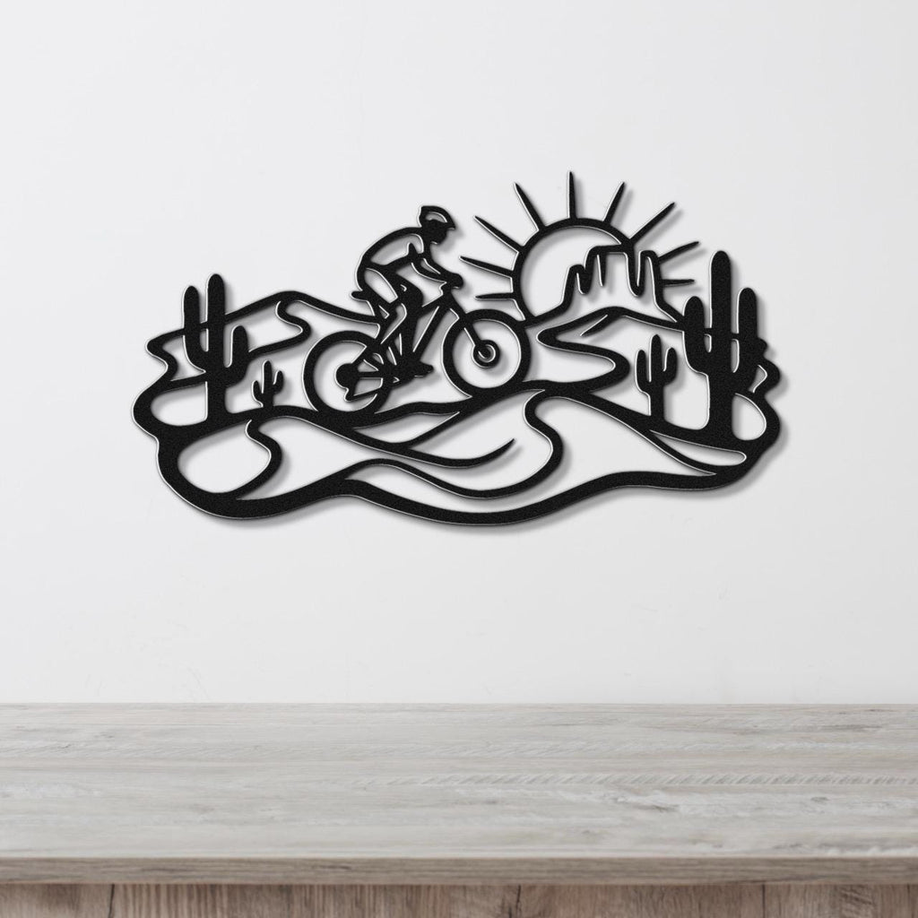 Black metal wall art featuring a cyclist silhouette on a desert trail with cacti and a sunburst, mounted on a white wall above a wooden surface.