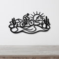 Black metal wall art featuring a cyclist silhouette on a desert trail with cacti and a sunburst, mounted on a white wall above a wooden surface.