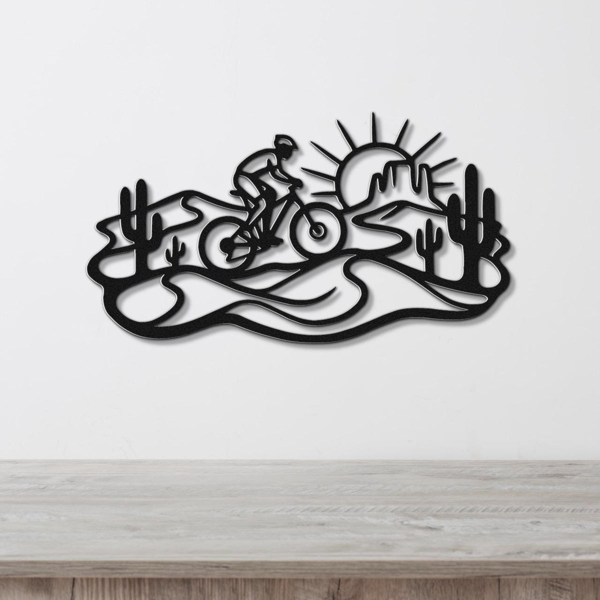 Black metal wall art featuring a cyclist silhouette on a desert trail with cacti and a sunburst, mounted on a white wall above a wooden surface.