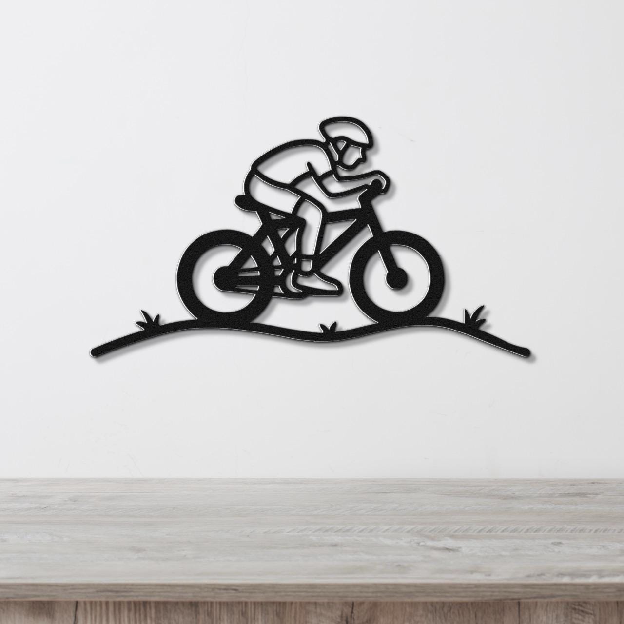 Black metal wall art featuring a minimalist cyclist silhouette in motion, mounted on a white wall above a wooden surface.