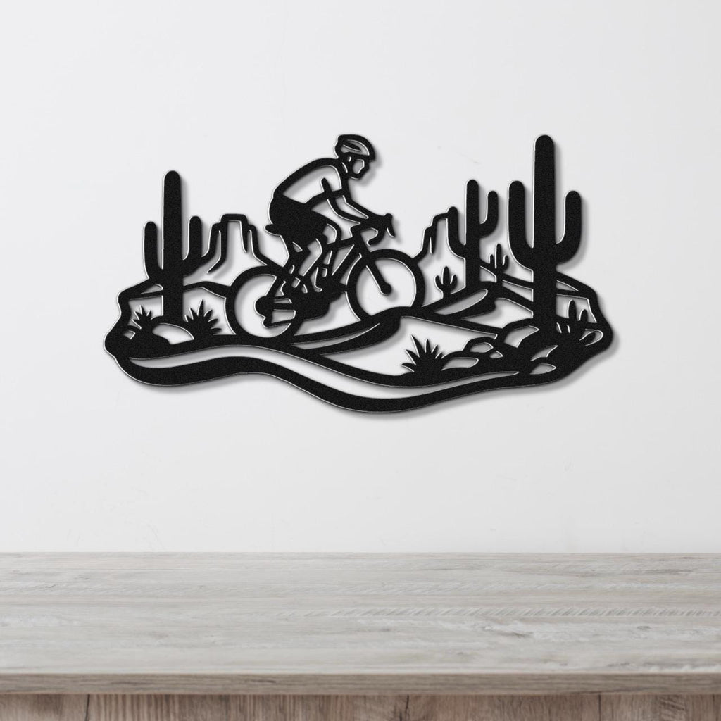Black metal wall art featuring a cyclist silhouette on a mountain bike through a Sonoran desert landscape with saguaro cacti and rocky terrain, mounted on a white wall above a wooden surface.