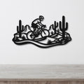 Black metal wall art featuring a cyclist silhouette on a mountain bike through a Sonoran desert landscape with saguaro cacti and rocky terrain, mounted on a white wall above a wooden surface.