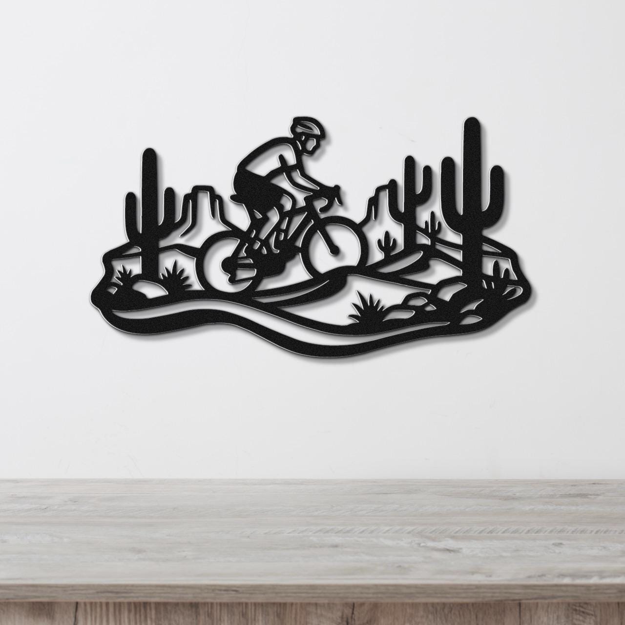 Black metal wall art featuring a cyclist silhouette on a mountain bike through a Sonoran desert landscape with saguaro cacti and rocky terrain, mounted on a white wall above a wooden surface.