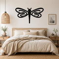 Black metal dragonfly wall art with flowing wing cutouts, mounted above a wooden bed in a neutral bedroom.