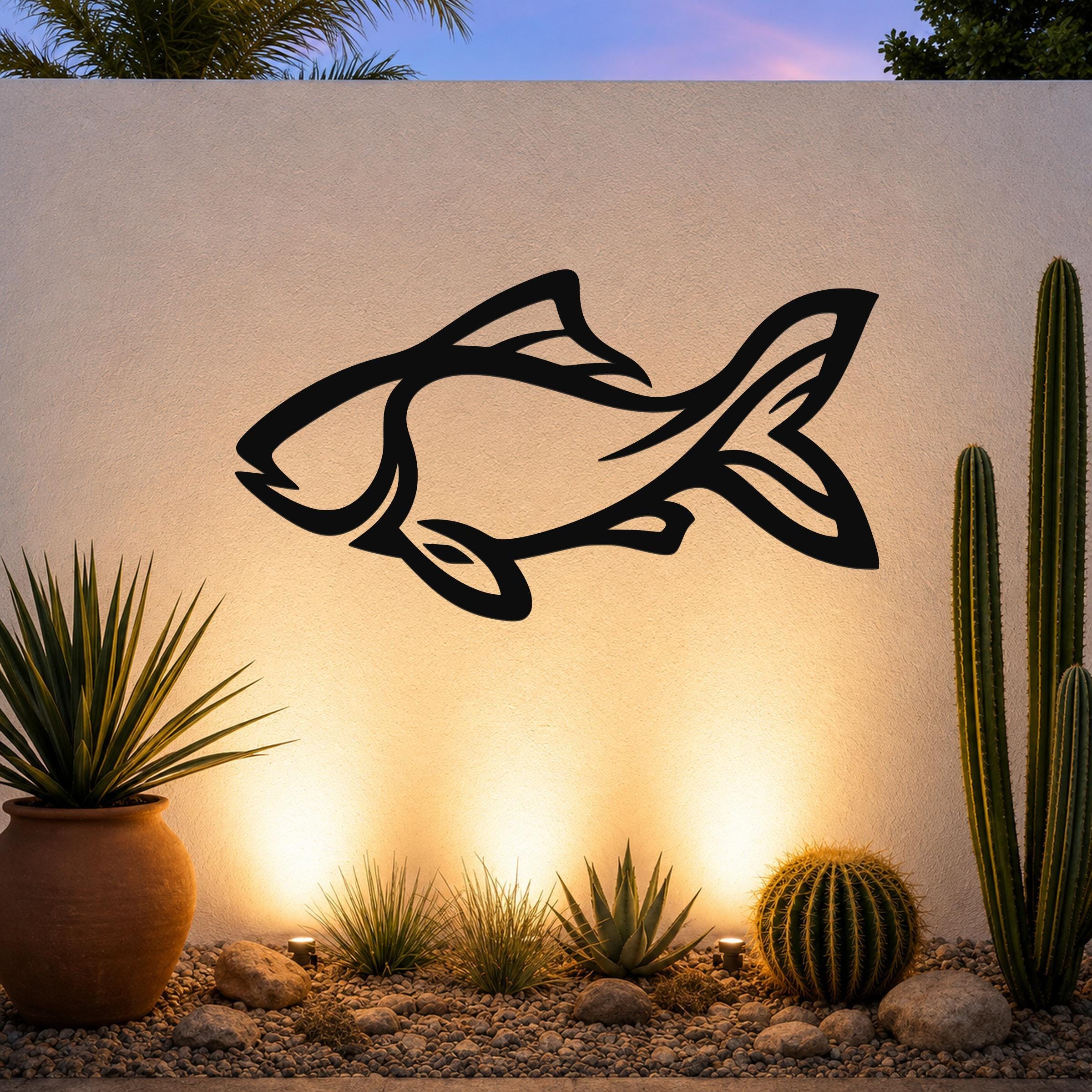 Black metal fish wall art with internal cutout details, mounted on a textured outdoor wall above a desert garden with cacti and warm uplighting.