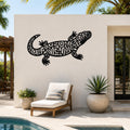 Black metal wall art of a Gila monster with intricate cutout patterns, mounted on an outdoor patio wall above a lounge chair near a pool.