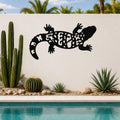 Black metal Gila monster wall art with intricate cutout patterns, mounted on a white outdoor wall beside a pool and desert plants.