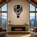 Black metal hot air balloon wall art with swirling internal cutouts, mounted above a fireplace in a rustic living room with large windows overlooking snowy mountains.
