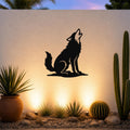 Black metal coyote wall art in a howling silhouette, mounted on an outdoor wall above a desert garden with cacti and warm uplighting.