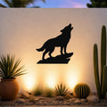 Black metal coyote wall art silhouette howling on a rock, mounted on an outdoor wall above a desert garden with cacti and warm uplighting.