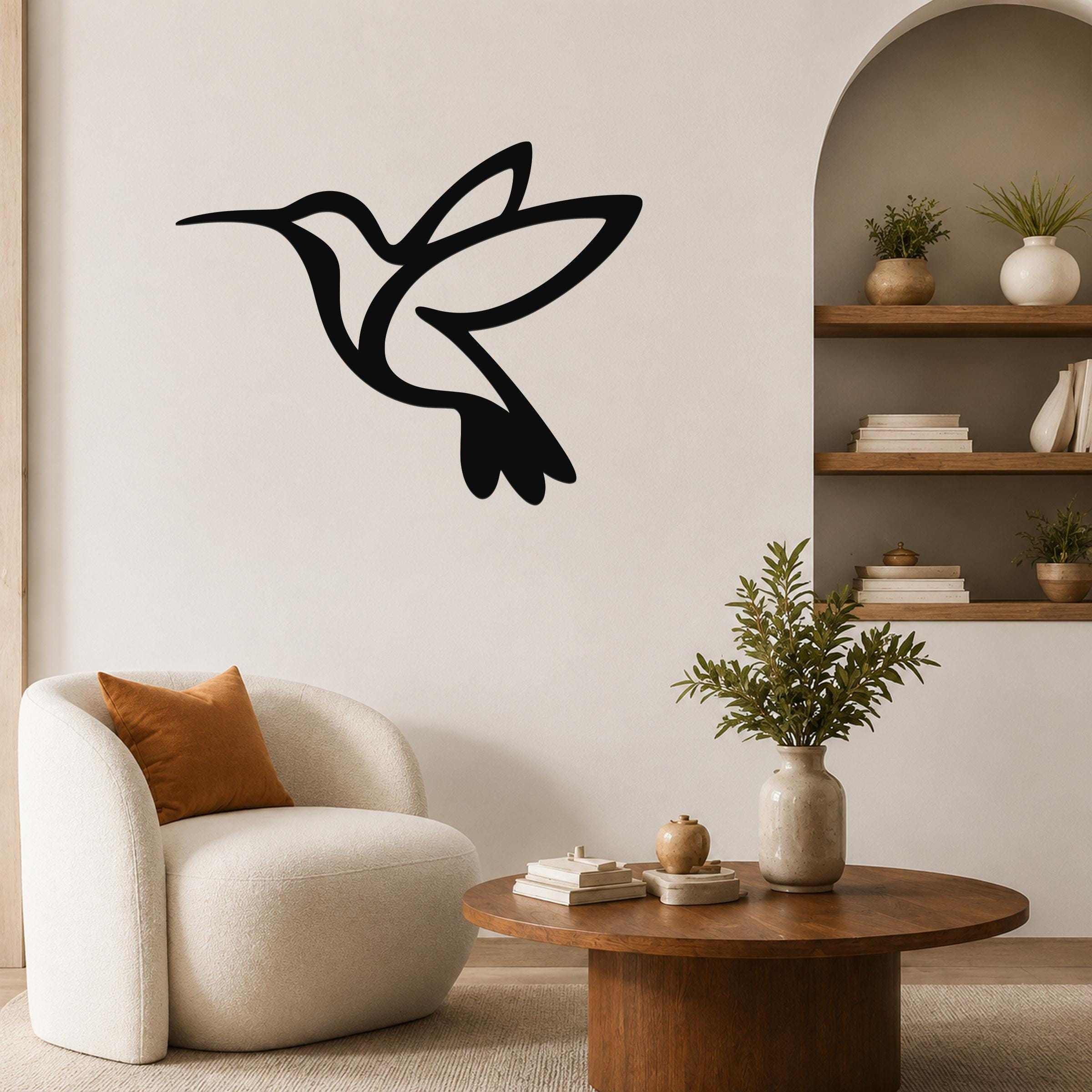 Black metal hummingbird wall art with a silhouette design, mounted on a living room wall above a white armchair.
