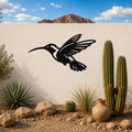 Black metal hummingbird wall art with an airy silhouette design, mounted on a white outdoor wall above a desert garden with cacti and rocks.