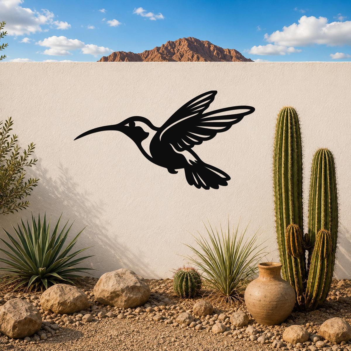 Black metal hummingbird wall art with an airy silhouette design, mounted on a white outdoor wall above a desert garden with cacti and rocks.