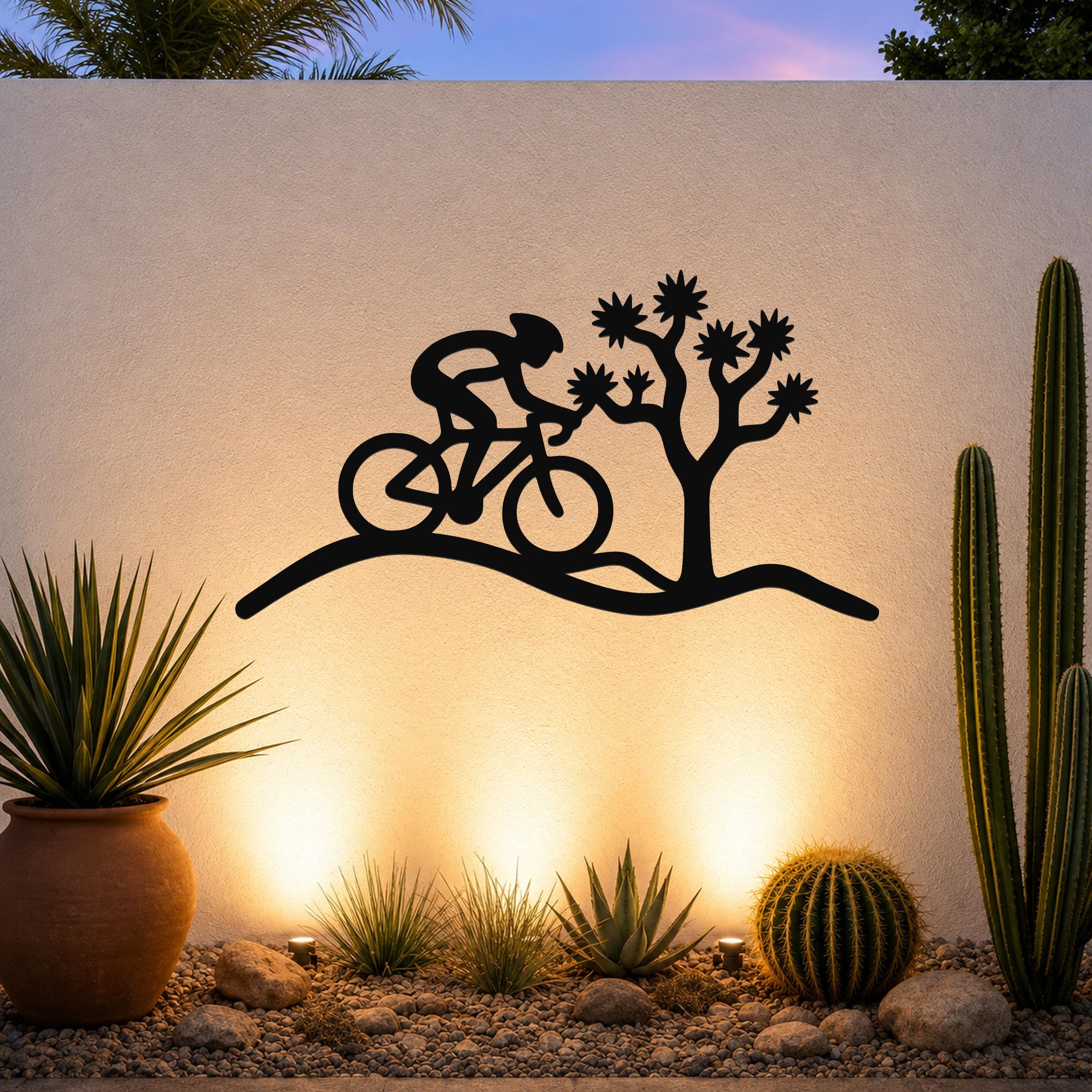 Black metal wall art silhouette of a cyclist on a desert trail with a Joshua tree, mounted on an outdoor wall above a gravel garden with cacti and warm uplighting.
