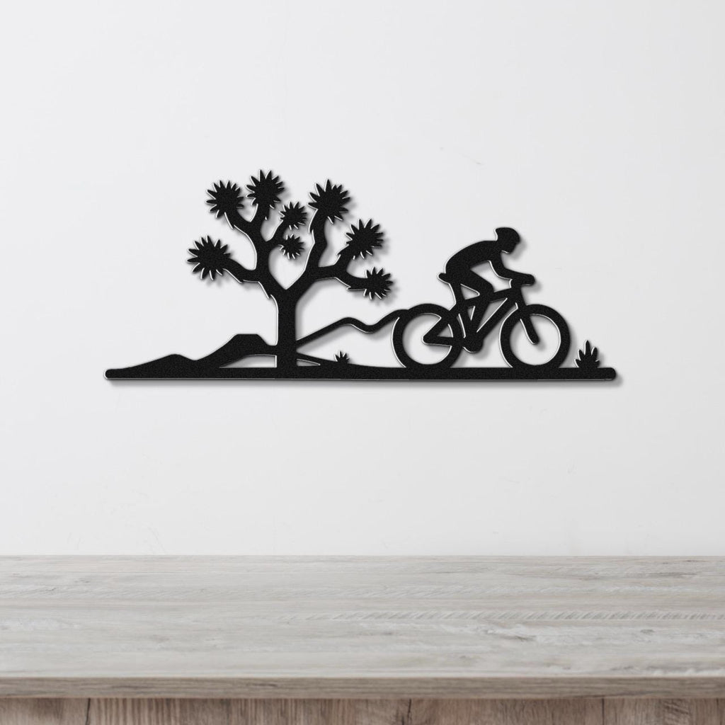 Black metal wall art silhouette of a cyclist on a mountain bike riding past a Joshua tree, mounted on a white wall above a wooden surface.