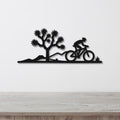 Black metal wall art silhouette of a cyclist on a mountain bike riding past a Joshua tree, mounted on a white wall above a wooden surface.