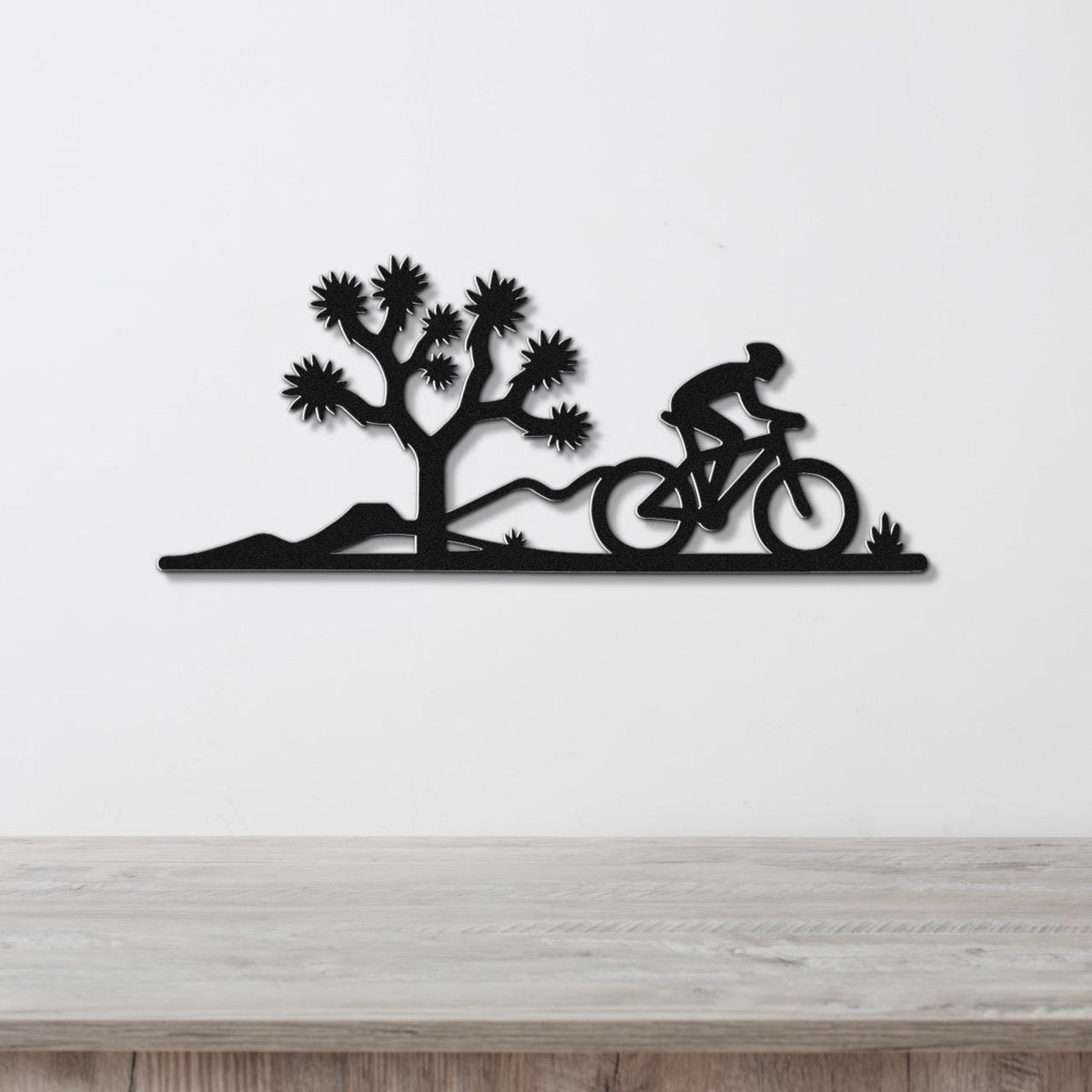 Black metal wall art silhouette of a cyclist on a mountain bike riding past a Joshua tree, mounted on a white wall above a wooden surface.