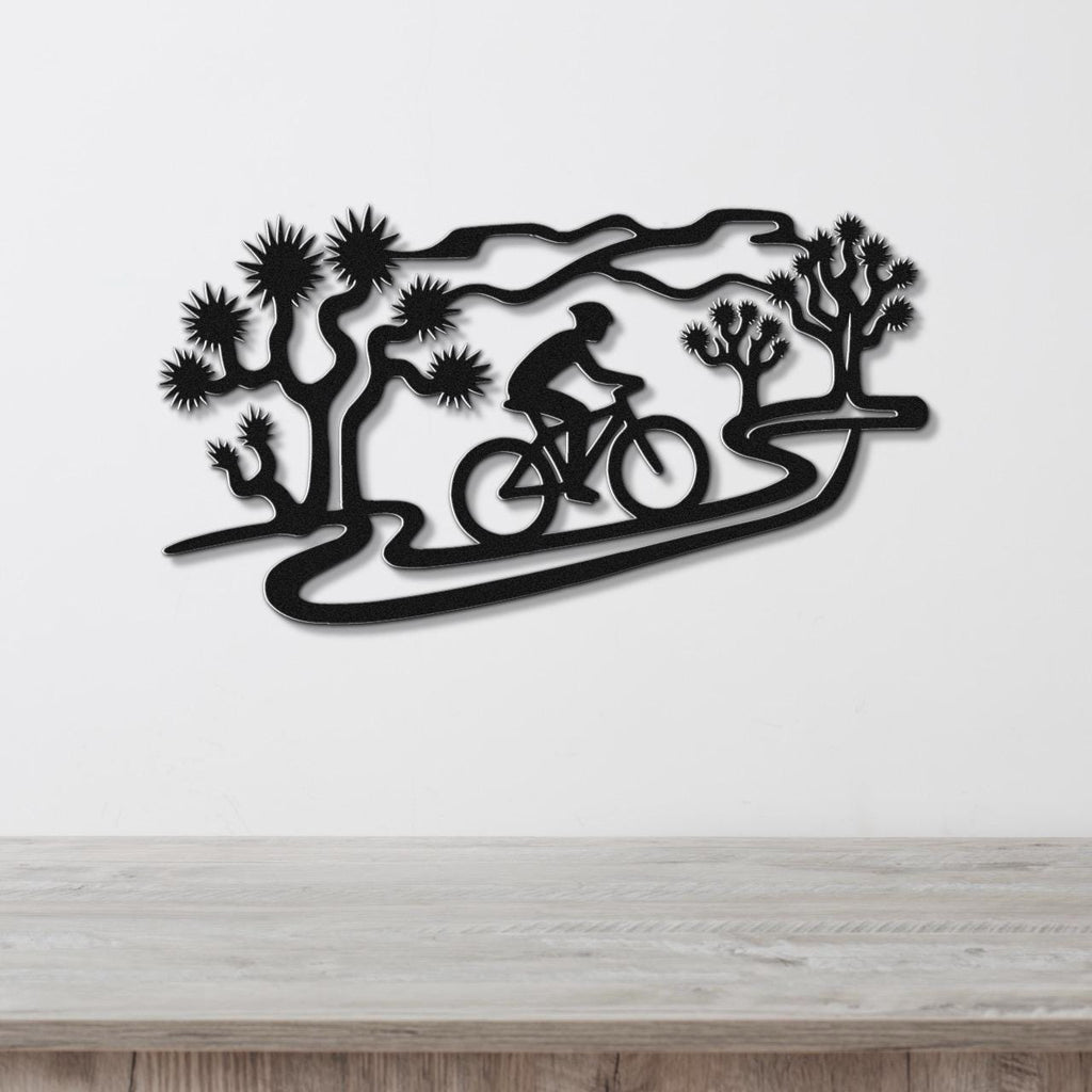 Black metal wall art featuring a cyclist silhouette on a winding path surrounded by Joshua trees, mounted on a white wall above a wooden surface.