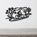 Black metal wall art featuring a cyclist silhouette on a winding path surrounded by Joshua trees, mounted on a white wall above a wooden surface.
