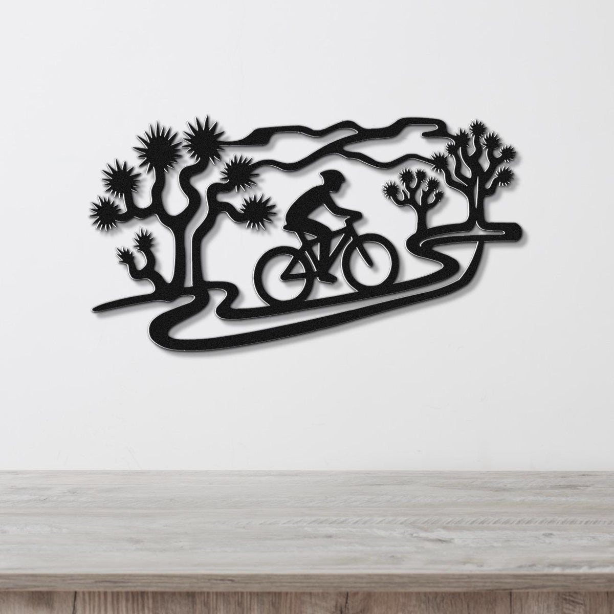 Black metal wall art featuring a cyclist silhouette on a winding path surrounded by Joshua trees, mounted on a white wall above a wooden surface.