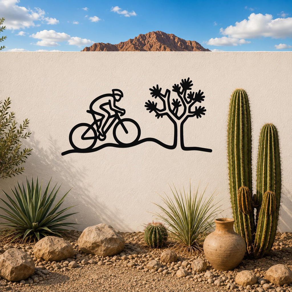 Black metal wall art silhouette of a cyclist and Joshua tree, mounted on a white outdoor wall above a desert garden with cacti and rocks.