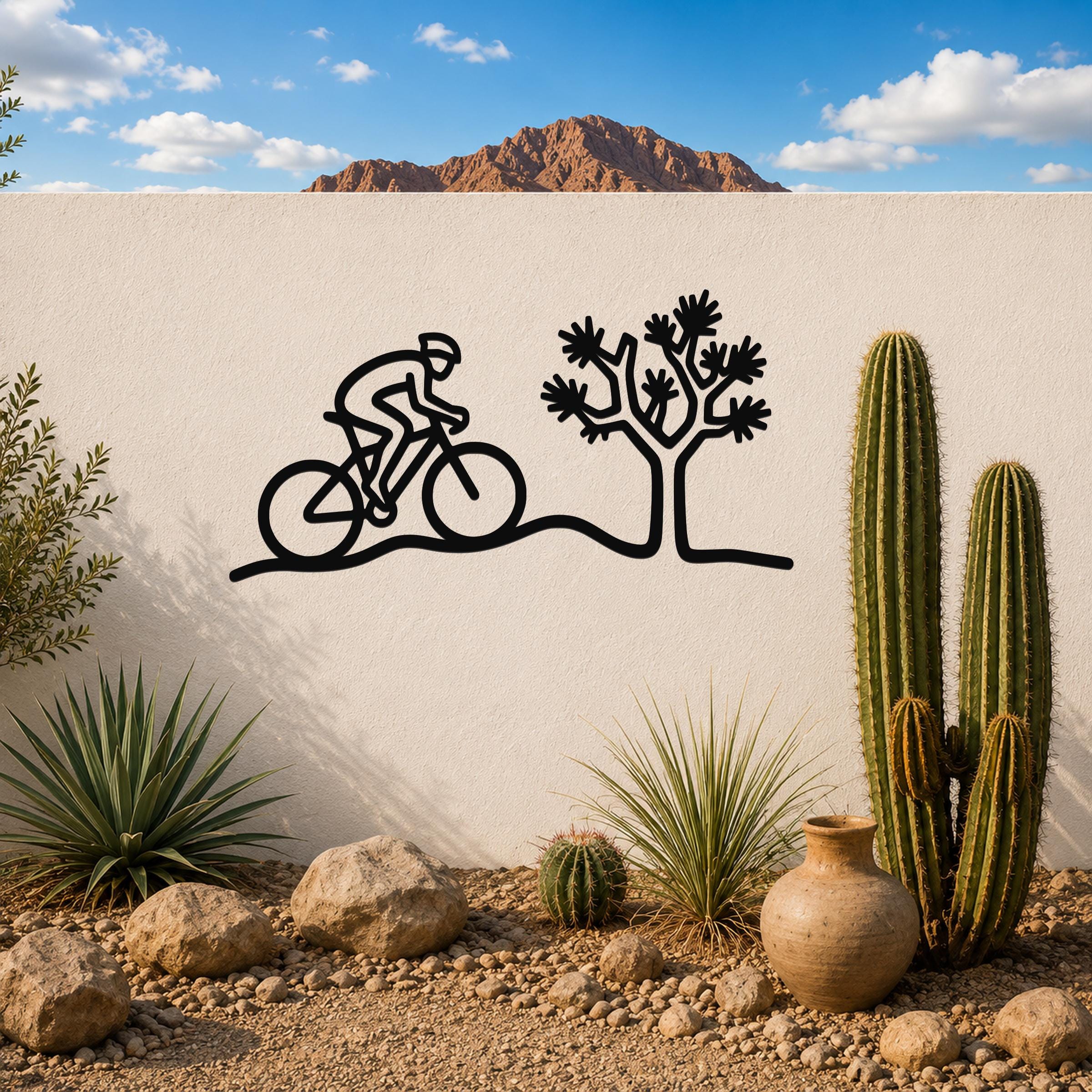 Black metal wall art silhouette of a cyclist and Joshua tree, mounted on a white outdoor wall above a desert garden with cacti and rocks.