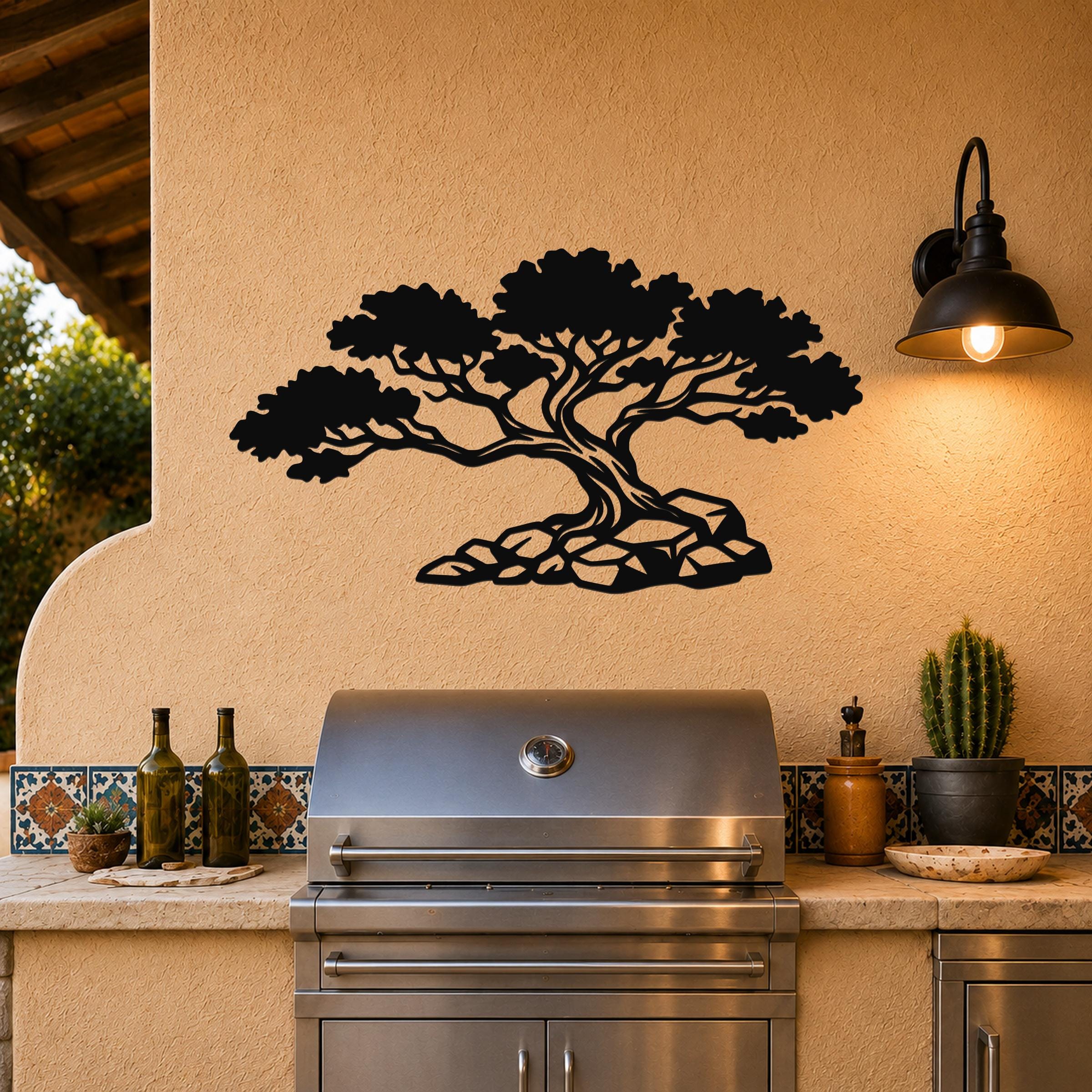 Black metal wall art cutout of a juniper tree with rocky base, mounted on a stucco wall above a stainless steel grill in an outdoor kitchen.