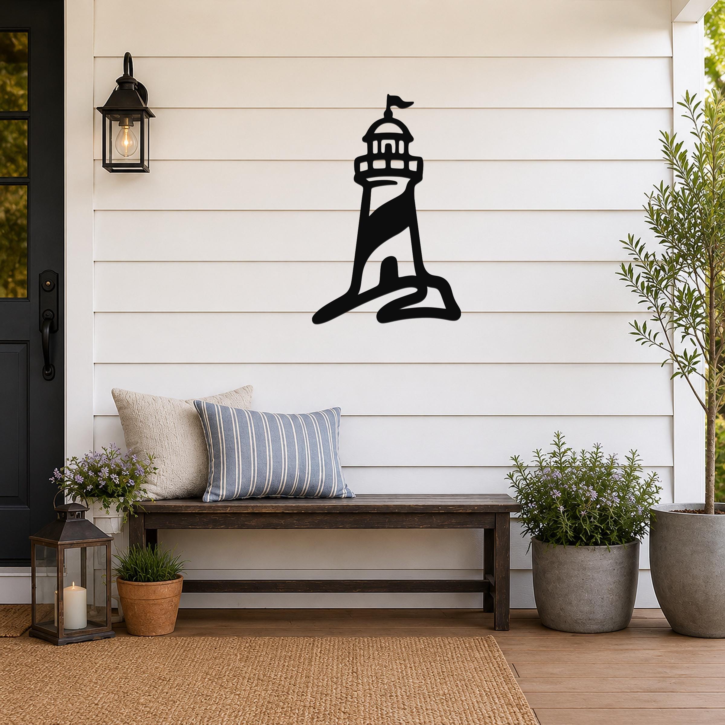 Black metal lighthouse wall art with a silhouette design, mounted on a white shiplap wall above a wooden bench on a porch.