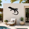 Black metal lizard wall art with internal cutout details, mounted on a light-colored outdoor wall beside a pool and lounge chair.