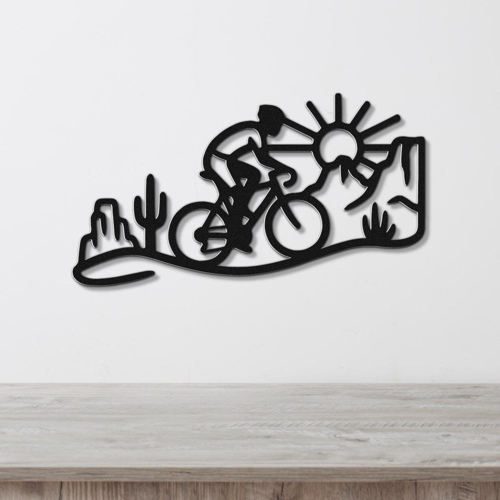 Black metal wall art silhouette of a cyclist in a desert scene with cacti and a sun, mounted on a white wall above a wooden counter.