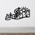 Black metal wall art silhouette of a cyclist in a desert scene with cacti and a sun, mounted on a white wall above a wooden counter.