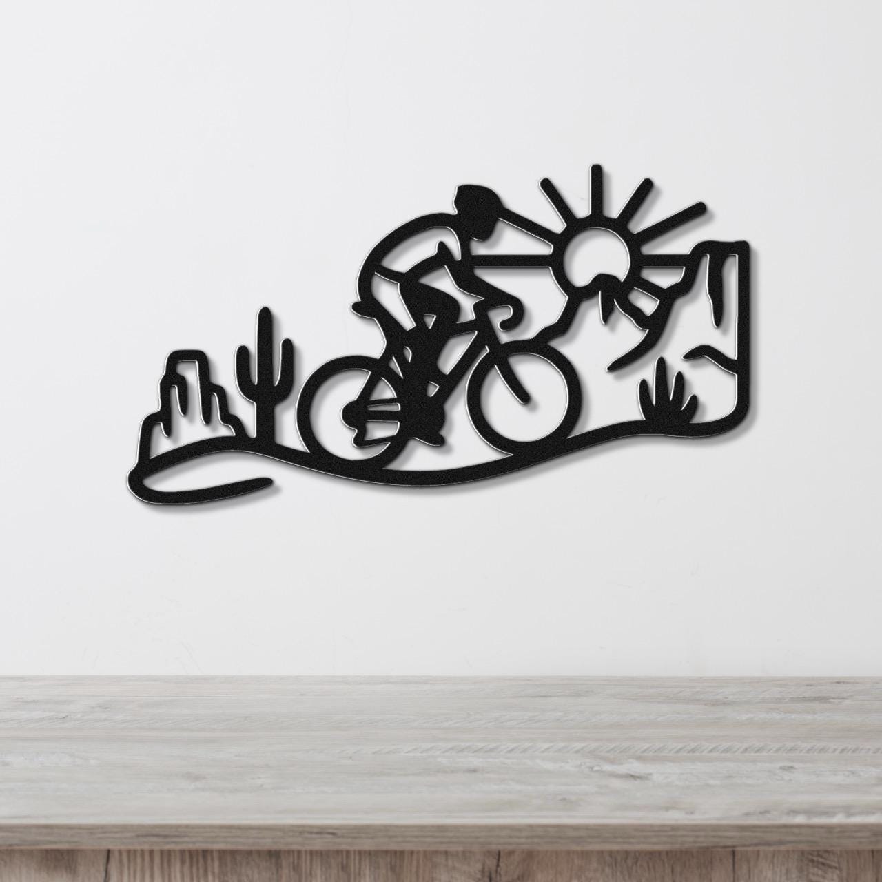 Black metal wall art silhouette of a cyclist in a desert scene with cacti and a sun, mounted on a white wall above a wooden counter.