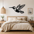Black metal hummingbird wall art with open wings and internal cutout details, mounted above a wooden bed in a neutral bedroom.