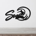 Black metal wall art featuring a surfer silhouette riding a wave, with the word "Surf" in cursive script, mounted on a white wall above a light wood surface.