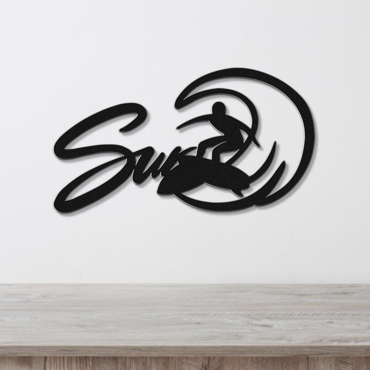 Black metal wall art featuring a surfer silhouette riding a wave, with the word "Surf" in cursive script, mounted on a white wall above a light wood surface.