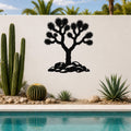 Black metal wall art silhouette of a Joshua tree with spiky branches and rocks at its base, mounted on a white outdoor wall above a poolside garden.