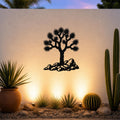 Black metal wall art featuring a Joshua tree silhouette with spiky branches and rocks at its base, mounted on a light-colored outdoor wall above a desert garden with cacti and warm uplighting.